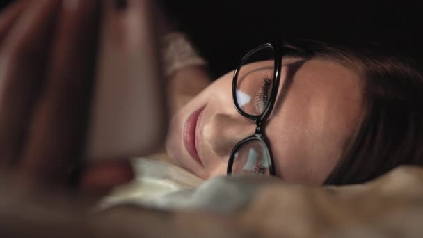 Donna con gli occhiali guarda smartphone e si trova sul letto, sorride sul viso — Video Stock