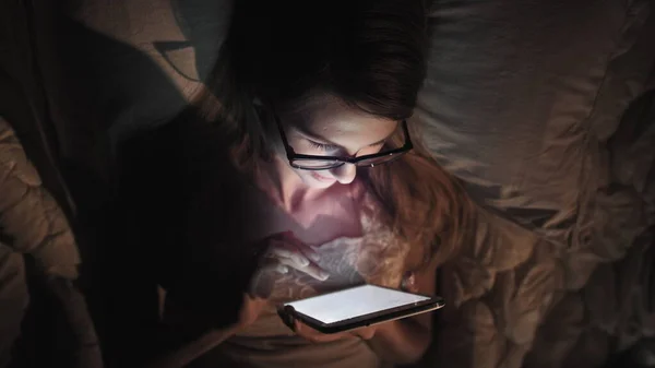 Una mujer con gafas mira un teléfono inteligente y se acuesta en la cama, jugueteando con los mensajes, buscando en Internet, vista superior — Foto de Stock
