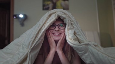 Kadın evde komedi izlemekten, kız battaniyenin altında yatmaktan zevk alıyor.