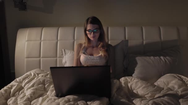 Młoda dobrze wyglądająca dziewczyna w okularach pisząca i pracująca na laptopie jako wolny strzelec siedząc w domu w przytulnym ciemnym salonie z kotem. Wieczorem. Zostań w domu. — Wideo stockowe