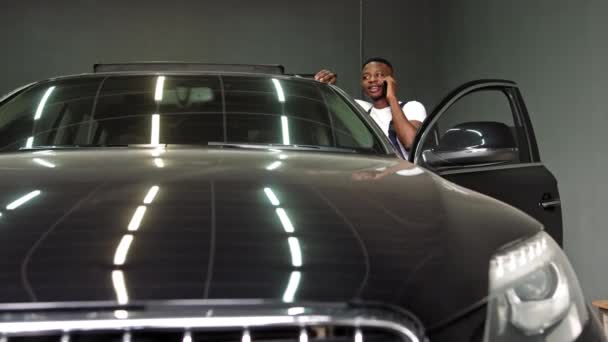 Młody Afroamerykanin rozmawiający przez telefon w pobliżu drogiego czarnego samochodu w garażu, portret w zwolnionym tempie — Wideo stockowe