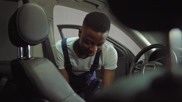 Retrato de un joven afroamericano, un estudiante que está vestido con ropa de trabajo especial, gana dinero lavando autos caros, un tipo alegre mira a la cámara y limpia la silla de auto — Vídeo de stock