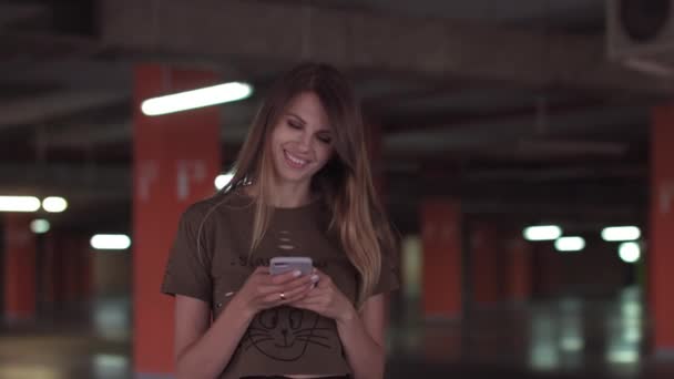 Zrelaksowana młoda kobieta korzystająca ze smartfona surfującego w mediach społecznościowych, sprawdzająca wiadomości, grająca w gry mobilne lub SMS-y stojąca na parkingu supermarketu. — Wideo stockowe