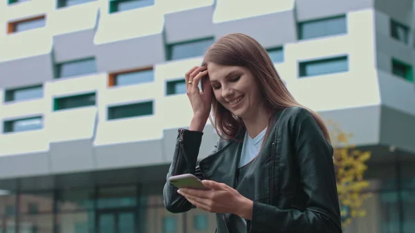 Jeune fille attrayante avec de longs cheveux bruns marchant dans la rue, ayant le téléphone portable dans ses mains. Dactylographier des messages et sourire lors de l'envoi. Avoir des vêtements à la mode. — Photo