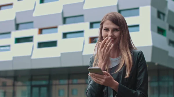 Ung, attraktiv pike med langt hår som går i gaten og har mobiltelefon i hendene. Maskinskriving og smil mens du sender. Å ha fasjonable klær. – stockfoto