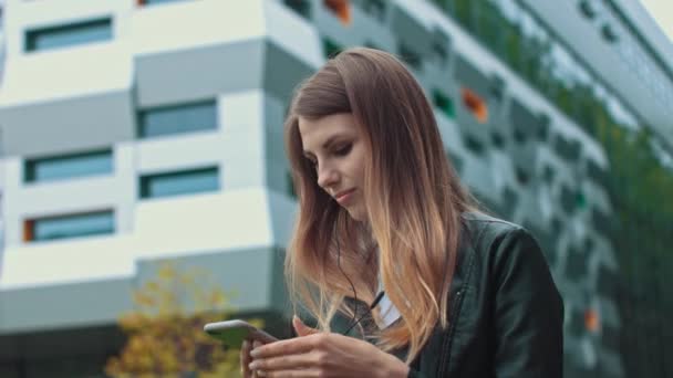 Close up of young girl, woman, attractive smiling young woman in modern leather clothes putting ears on headphones and having fun listening to music while walking around the city. After work on the — Stock Video
