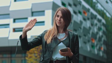 Uzun kahverengi saçlı, telefonuna bakıp gülümseyen güzel bir kız. Arka plandaki devasa sanayi binası. Yeşil çalılar ve ağaçlar. Güzel kıyafetler. Doğal makyaj..