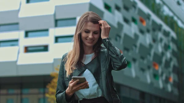 Belle fille avec de longs cheveux bruns En regardant son téléphone Dactylographier et sourire. Immense bâtiment industriel à l'arrière-plan. Arbres et buissons verts. Des vêtements intelligents. Maquillage naturel. — Photo