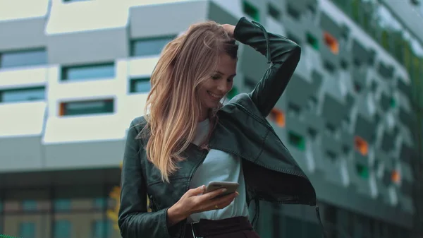 Belle fille avec de longs cheveux bruns En regardant son téléphone Dactylographier et sourire. Immense bâtiment industriel à l'arrière-plan. Arbres et buissons verts. Des vêtements intelligents. Maquillage naturel. — Photo