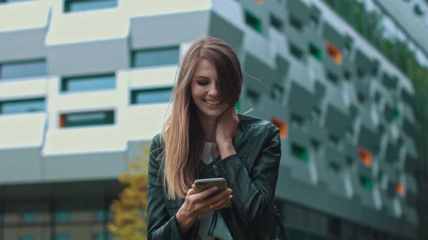 Pen jente med langt, brunt hår som ser på telefonen sin. Enorm industribygning i bakgrunnen. Grønne busker og trær. Smarte klær. Naturlig sminke. – stockfoto