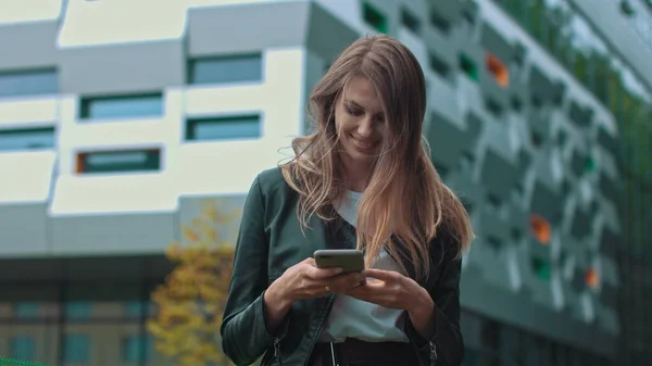 Pen jente med langt, brunt hår som ser på telefonen sin. Enorm industribygning i bakgrunnen. Grønne busker og trær. Smarte klær. Naturlig sminke. – stockfoto