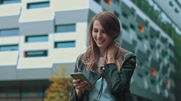 Pen jente med langt, brunt hår som ser på telefonen sin. Enorm industribygning i bakgrunnen. Grønne busker og trær. Smarte klær. Naturlig sminke. stockbilde