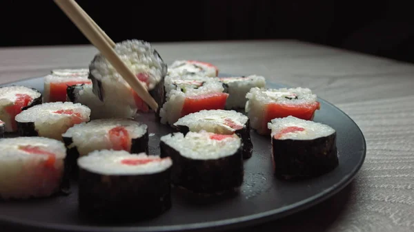 Golpe suave em um prato com comida japonesa. Mão feminina com pauzinhos leva sushi de uma bandeja. Sushi rola comida japonesa girada sobre fundo preto. Califórnia Sushi roll com atum, legumes — Fotografia de Stock