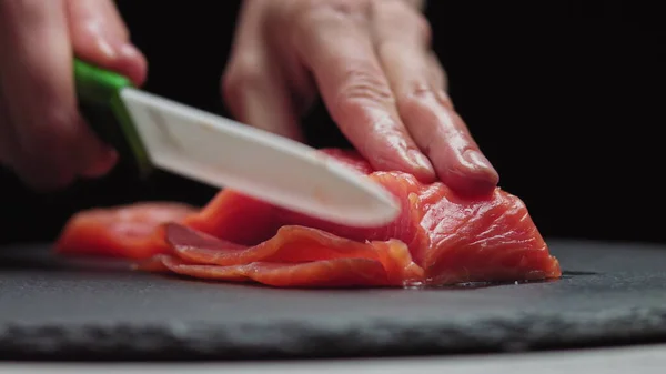 寿司職人寿司バーで新鮮なサーモンをスライスします。プロのキッチンで魚の切り身を切るシェフ。遅い動きで新鮮な魚のスライスをスライスシェフの手を閉じます.プロの男は赤魚を切断 — ストック写真