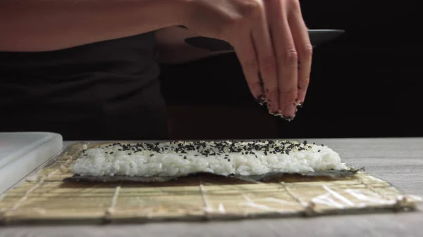 厨师们把芝麻种子撒到寿司里面.日本厨师戴着黑色手套在工作中准备用鲑鱼和鳄梨做寿司卷. — 图库照片
