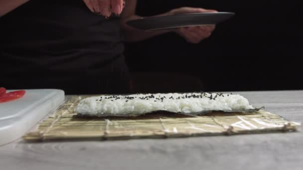 Chefs mãos polvilhe sementes de gergelim no sushi de dentro para fora. chef japonês em luvas pretas no trabalho preparando rolo de sushi com salmão e abacate. — Vídeo de Stock