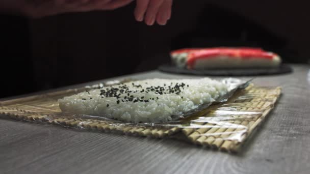 寿司の内側にゴマを振りかける料理人の手。黒毛和牛の日本人シェフが鮭とアボカドの巻き寿司の準備をしています. — ストック動画