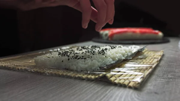 厨师们把芝麻种子撒到寿司里面.日本厨师戴着黑色手套在工作中准备用鲑鱼和鳄梨做寿司卷. — 图库照片