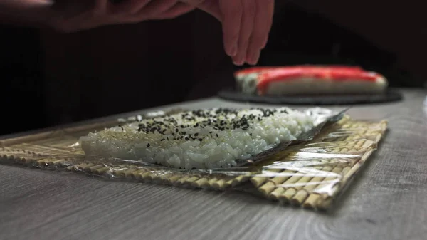 Chefs mãos polvilhe sementes de gergelim no sushi de dentro para fora. chef japonês em luvas pretas no trabalho preparando rolo de sushi com salmão e abacate. — Fotografia de Stock