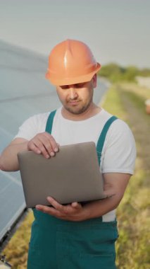 Sert şapkalı bir mühendis güneş panellerini incelemek için dizüstü bilgisayar kullanıyor. Dikey video.