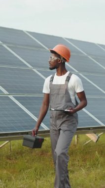 An African-American technician in safety gear inspects solar panels, holding a toolbox and clipboard, in a field.