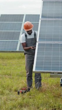 An African American technician in a hard hat inspects solar panels in a field, focusing on renewable energy.