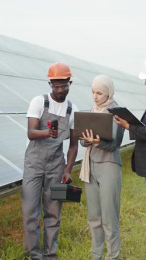 A technician in safety gear inspects solar panels at a solar farm, holding a toolbox. This is a vertical video.
