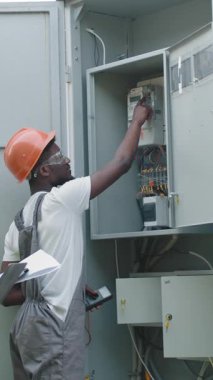 An electrician in a hard hat inspects an electrical panel, likely for maintenance or repairs. This vertical video captures the professional at work.