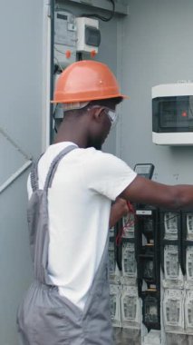 An electrician in a hard hat and safety glasses checks an electrical panel with a multimeter. Vertical video.