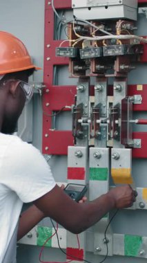 An electrician in a hard hat and safety glasses uses a multimeter to inspect an electrical panel, ensuring safety and functionality.