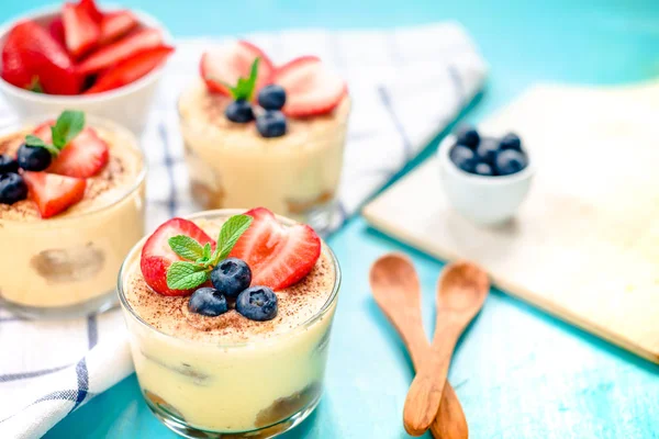 Homemade, exquisite dessert tiramisu in glasses decorated with strawberry, blueberry, mint on blue wooden table — Stock Photo, Image