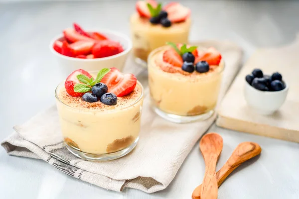 Homemade, exquisite dessert tiramisu in glasses decorated with strawberry, blueberry, mint on white wooden table, top view. — Stock Photo, Image