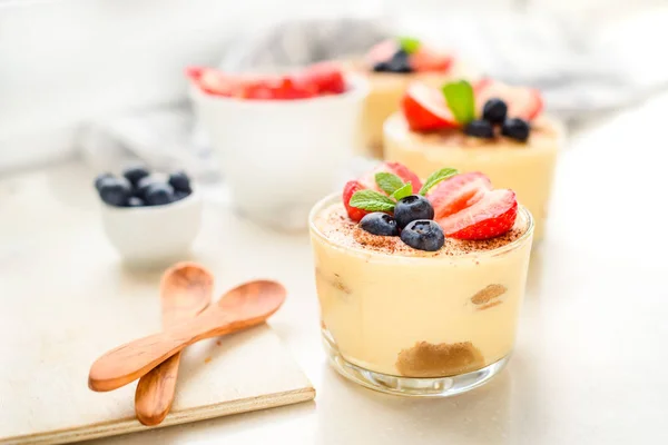 Homemade, exquisite dessert tiramisu in glasses decorated with strawberry, blueberry, mint on white wooden table, top view. — Stock Photo, Image