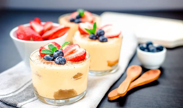 Homemade, exquisite dessert tiramisu in glasses decorated with strawberry, blueberry, mint on black wooden table — Stock Photo, Image