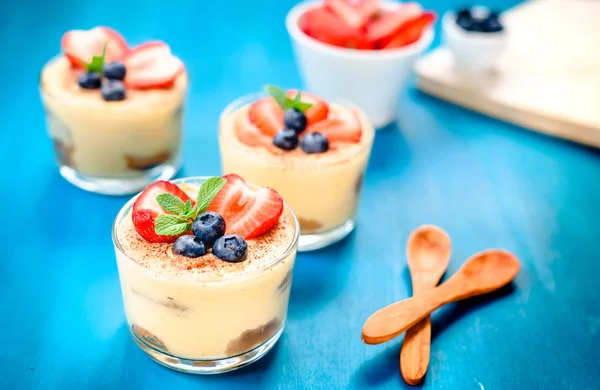 Homemade, exquisite dessert tiramisu in glasses decorated with strawberry, blueberry, mint on blue wooden table — Stock Photo, Image