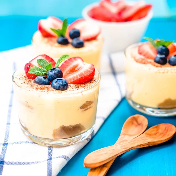 Homemade, exquisite dessert tiramisu in glasses decorated with strawberry, blueberry, mint on blue wooden table — Stock Photo, Image