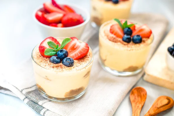 Homemade, exquisite dessert tiramisu in glasses decorated with strawberry, blueberry, mint on white wooden table — Stock Photo, Image