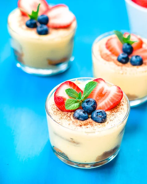 Homemade, exquisite dessert tiramisu in glasses decorated with strawberry, blueberry, mint on blue wooden table — Stock Photo, Image