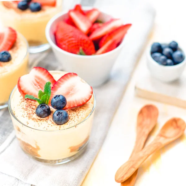 Homemade, exquisite dessert tiramisu in glasses decorated with strawberry, blueberry, mint on white wooden table. — Stock Photo, Image