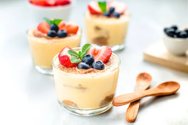 Homemade, exquisite dessert tiramisu in glasses decorated with strawberry, blueberry, mint on white wooden table — Stock Photo, Image