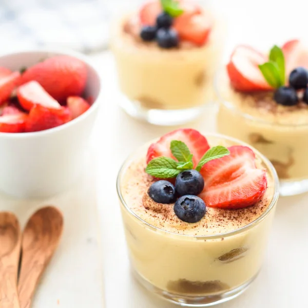 Homemade, exquisite dessert tiramisu in glasses decorated with strawberry, blueberry, mint on white wooden table — Stock Photo, Image