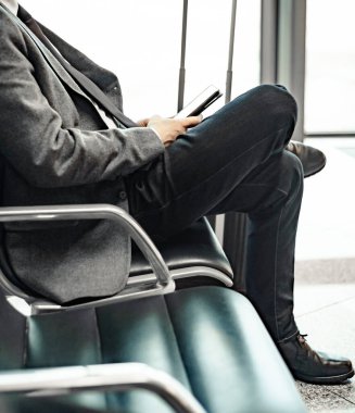 Adam havaalanında uçağını bekliyor, bavulun üstünde bacakları ve cep telefonuna bakıyor.