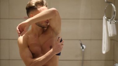 Genç adam banyoda, koltukaltlarına deodorant sıkıyor.