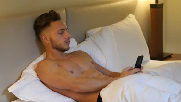 Hemdloser muskulöser junger Mann im Bett tippt auf Handy — Stockvideo