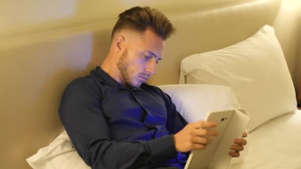 Красивый молодой человек в постели печатает на планшетном компьютере — стоковое видео