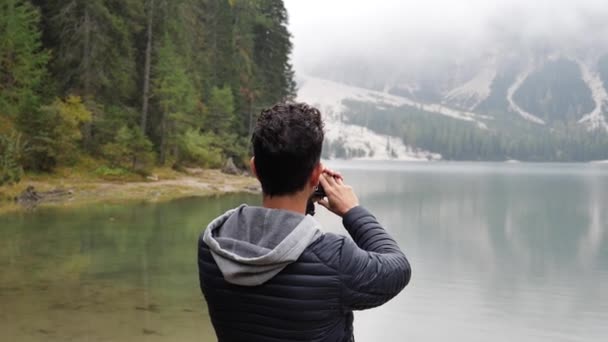 Молодой человек фотографирует или записывает видео с видеокамеры на озере — стоковое видео
