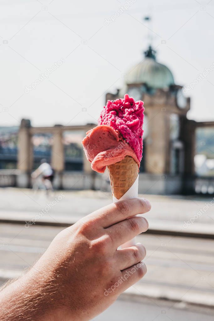 Bright pink ice-cream in a wafer cone held in man`s hand, blurred city on the background.