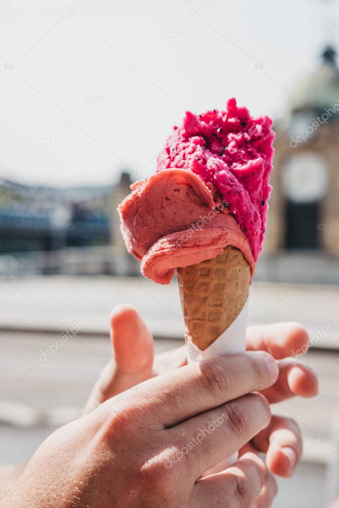 Bright pink ice-cream in a wafer cone held in man`s hand, blurred city on the background.