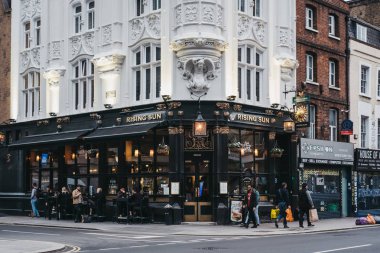 Londo'da bir Rising Sun pub önünde bir sokakta yürüyen insanlar