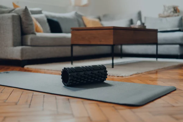 Sturdy black foam roller on a fitness mat, interior of a modern living room, selective shallow focus.